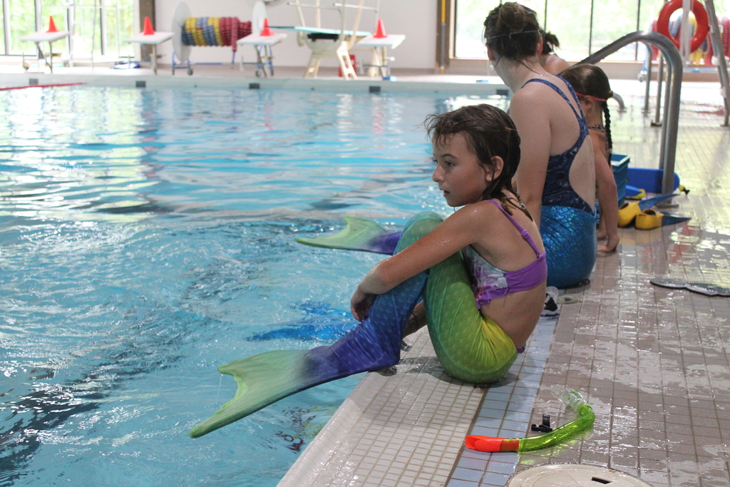 Child sits on edge of pool while wearing mermaid fin.
